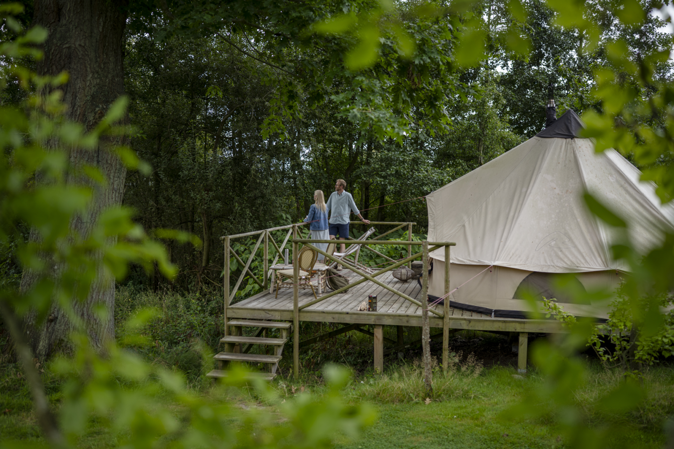 Par står foran hvidt glampingtelt på platform med træer i baggrunden