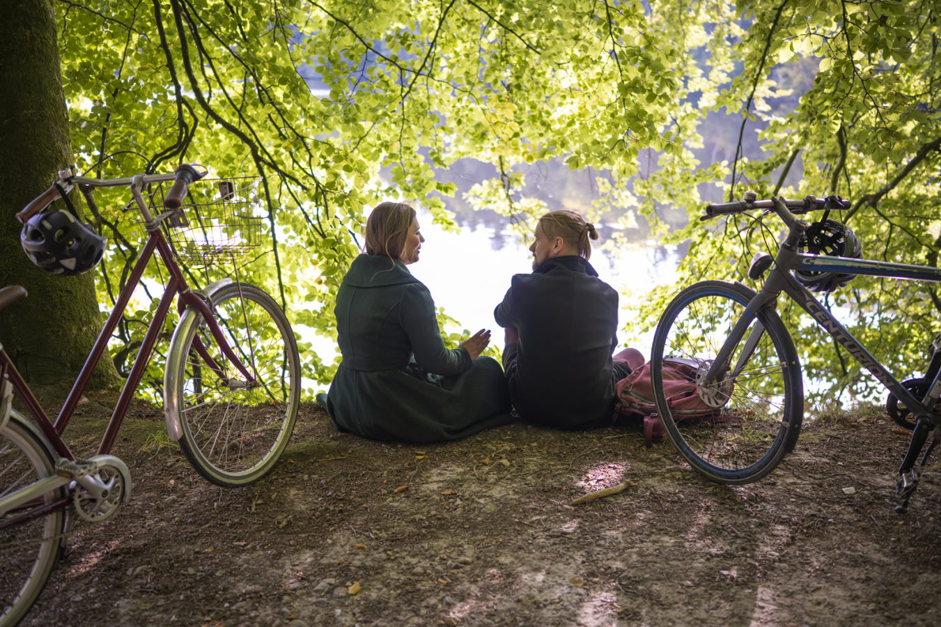 Par sidder i skovbrynet med ryggen til. En cykel er parkeret på hver side af dem.