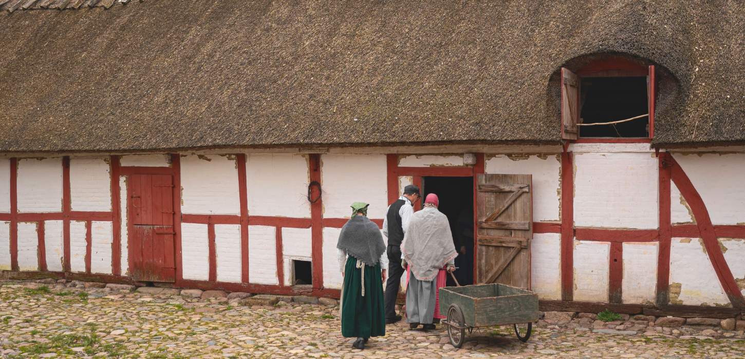Et par mennesker iklædt tøj fra tidligere århundreder er på vej ind i et rød og hvidt bindingsværkhus