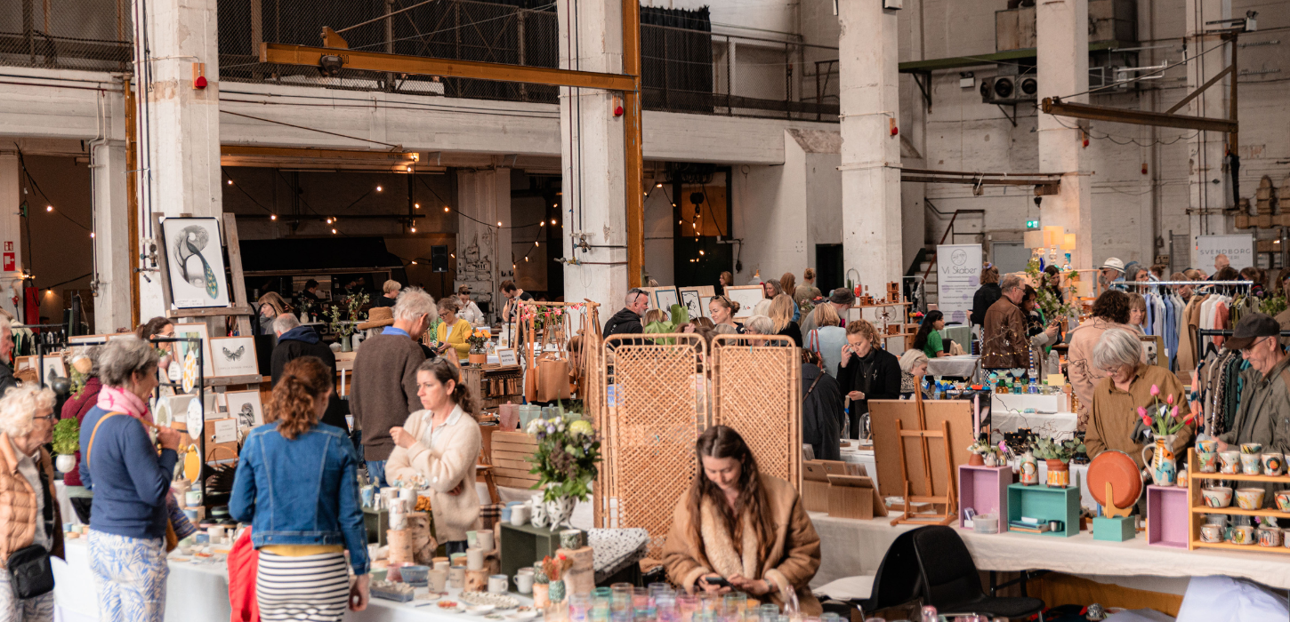 Et marked i en hal med højt til loftet er i fuld gang med mennesker, borde med varer og ting.