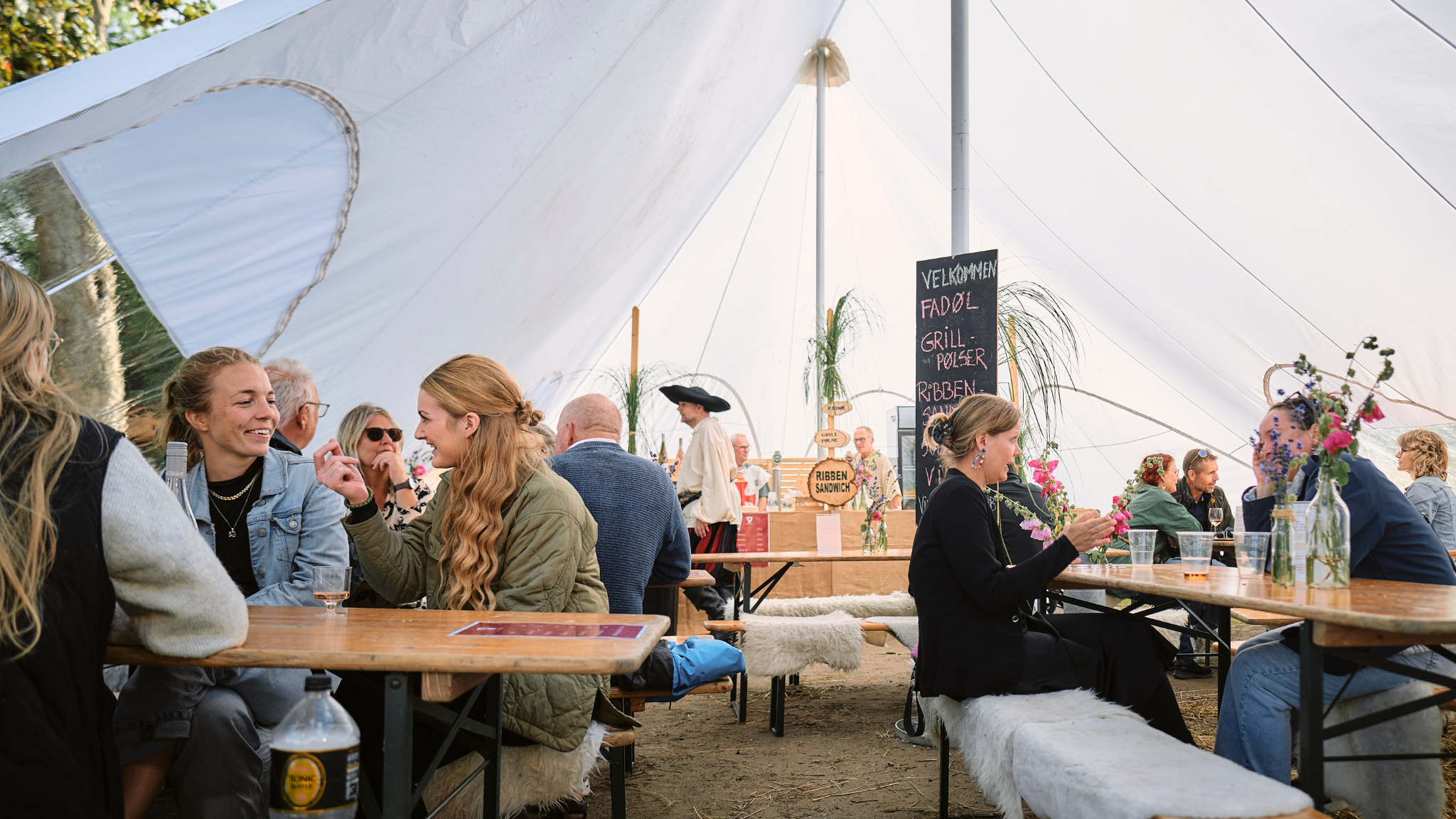 Festivalgæster sidder ved langborde i et hvidt telt, der er pyntet middelalderligt.