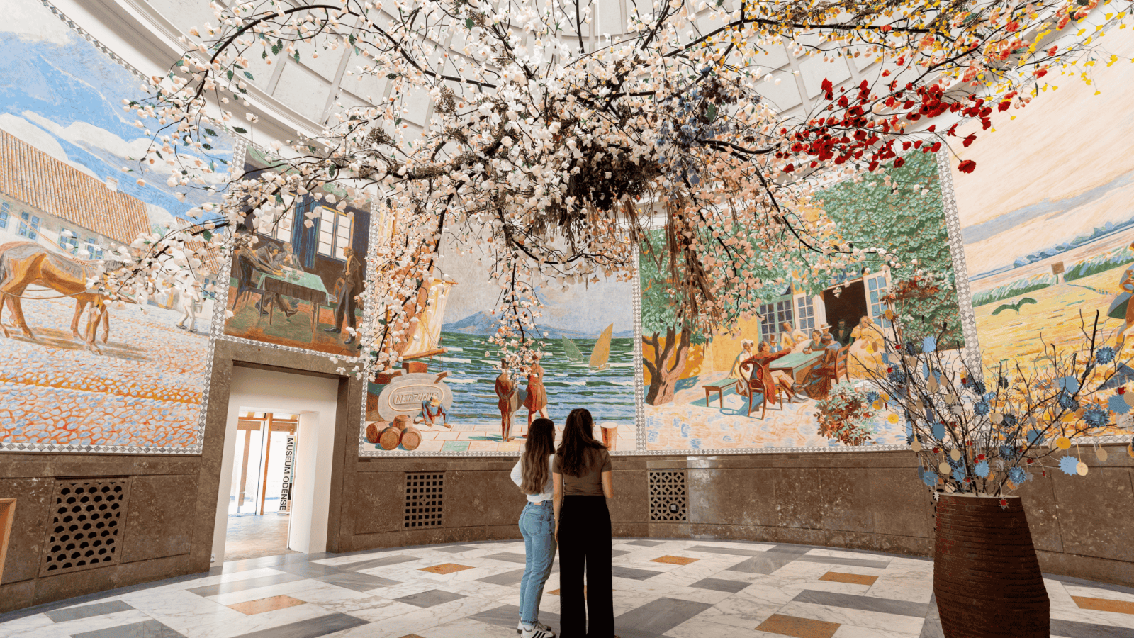 To piger står med ryggen til i en rund hal med papirsblomster hængende fra loftet