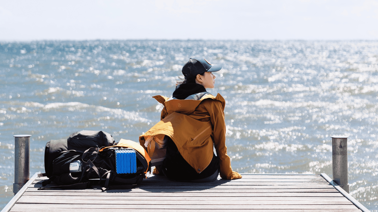 En kvindelig vandrer sidder iført regntøj på badebro med udsigt til himmel og hav
