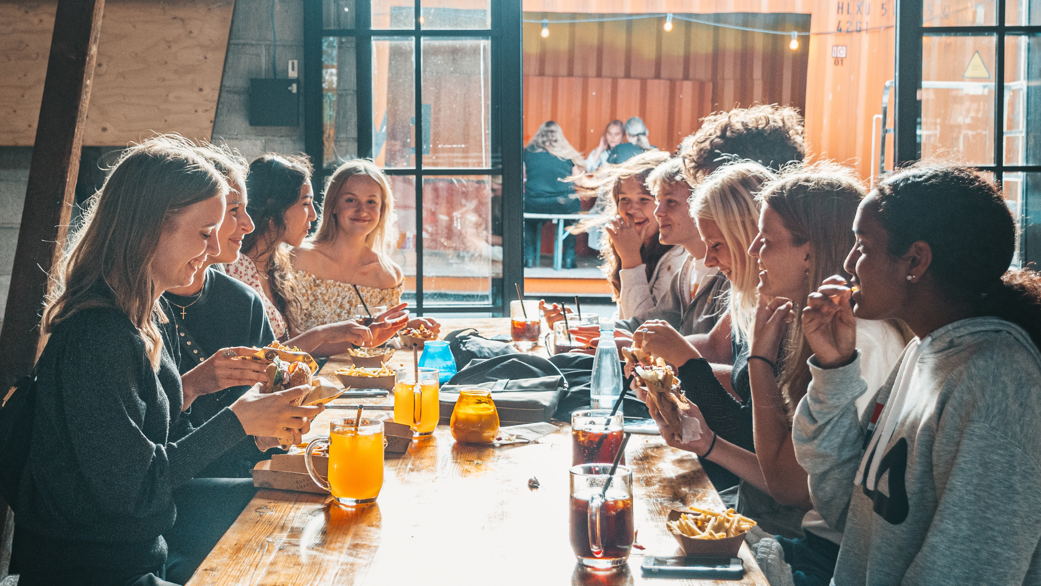 En gruppe yngre piger sidder og spiser streetfood langs et rustikt langbord 