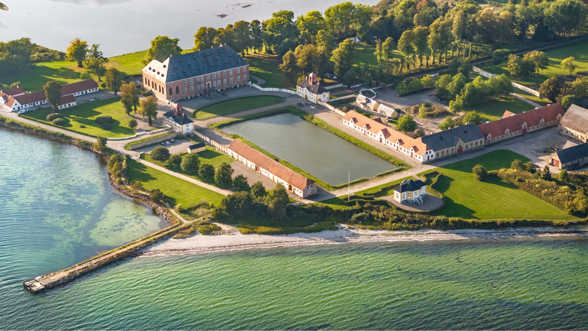 Et malerisk luftfoto af et slot med udsigt over en rolig sø, omgivet af træer og åbent landskab.