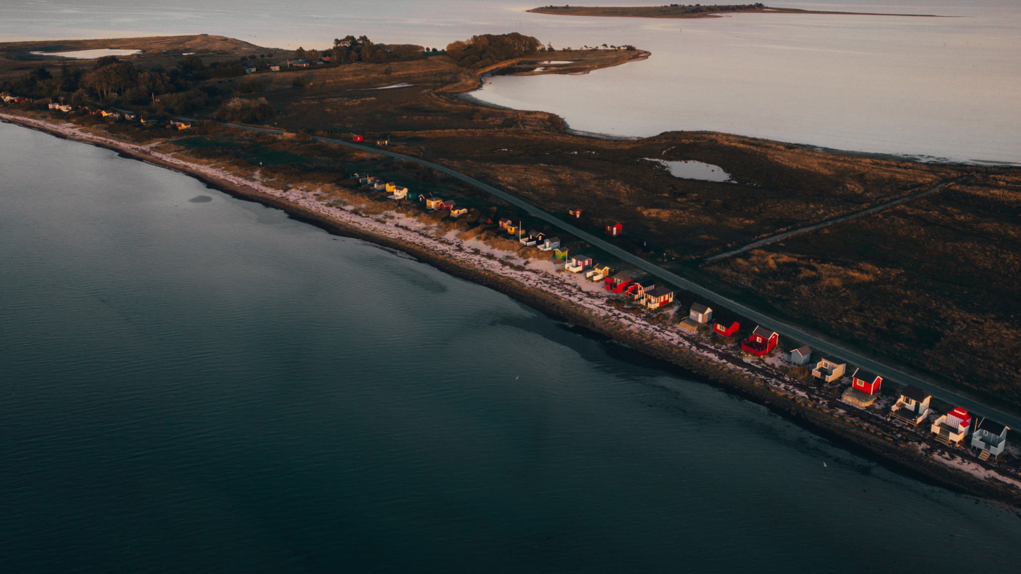 Luftfoto af en stribe farverige badehuse ved havet på en lang halvø, med bygninger og træer, der strækker sig langs kystlinjen.