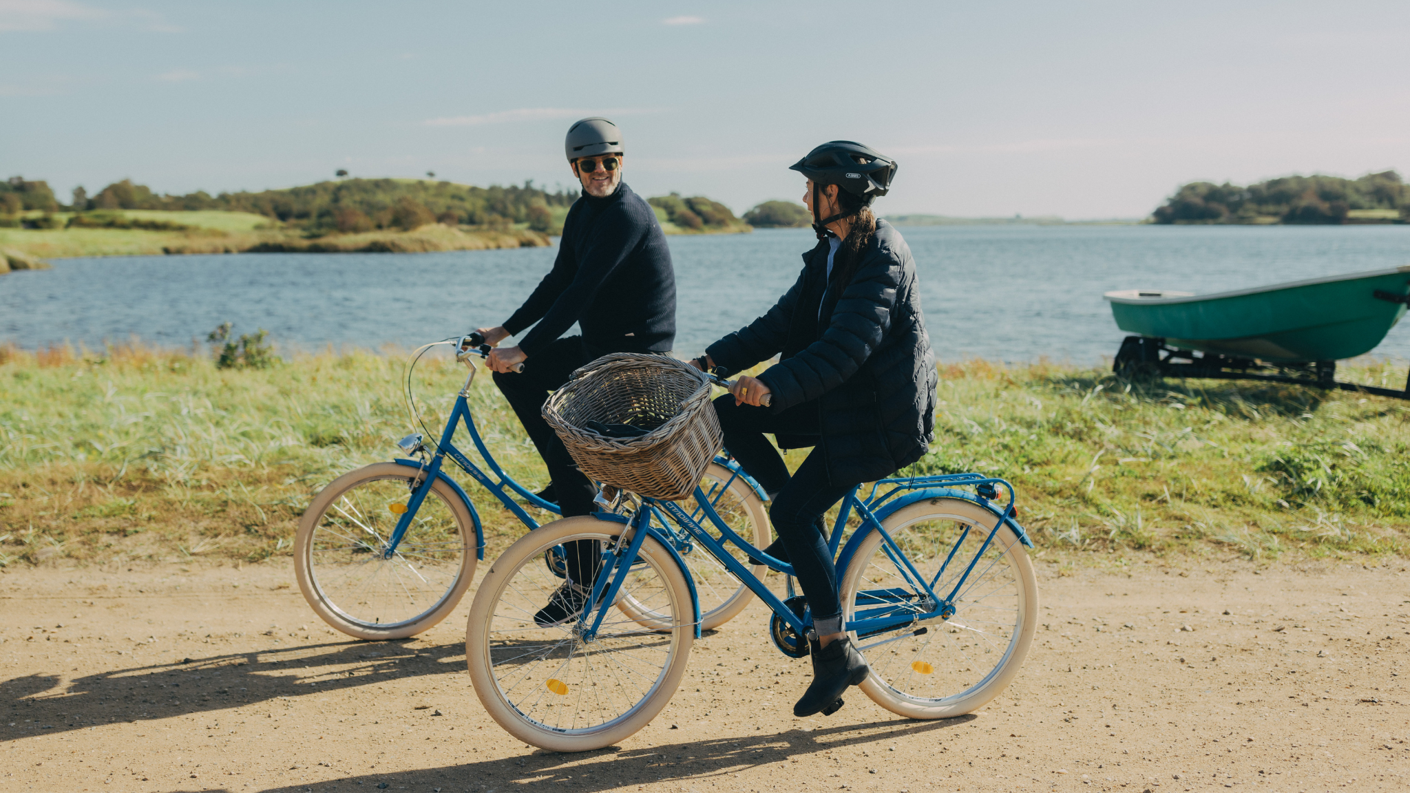 Et par i slutningen af fyrrerne cykler ved siden af hinanden på blå cykler på en grussti med havet i baggrunden.