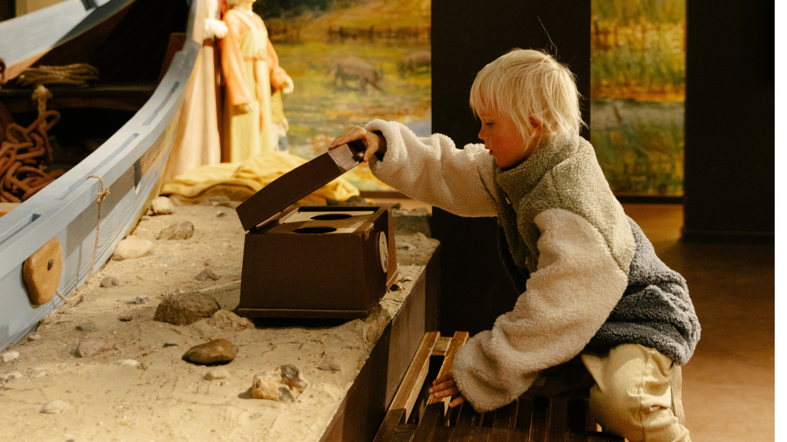 En lyshåret dreng i en fleecetrøje åbner en kasse i en udstilling på Vikingeskibsmuseet Ladby. 