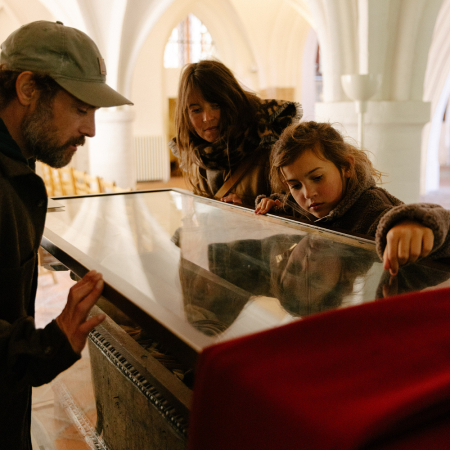 En mor, far og datter kigger ned i en glasmontre i kælderen af Odense Domkirke, hvor Knud den Helliges jordiske rester ligger i en montre. 