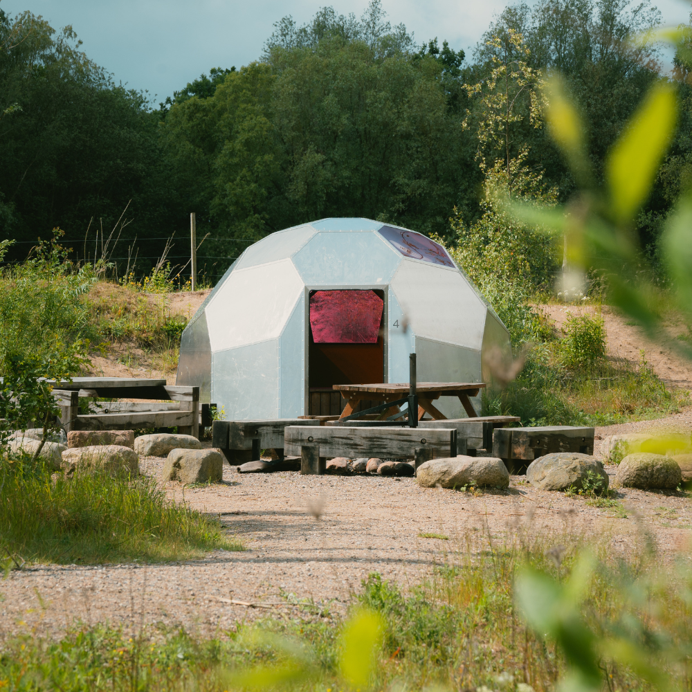 Et shelter formet som en kuppel i metal står ved en lejrplads omkranset af grønne træer og buske.