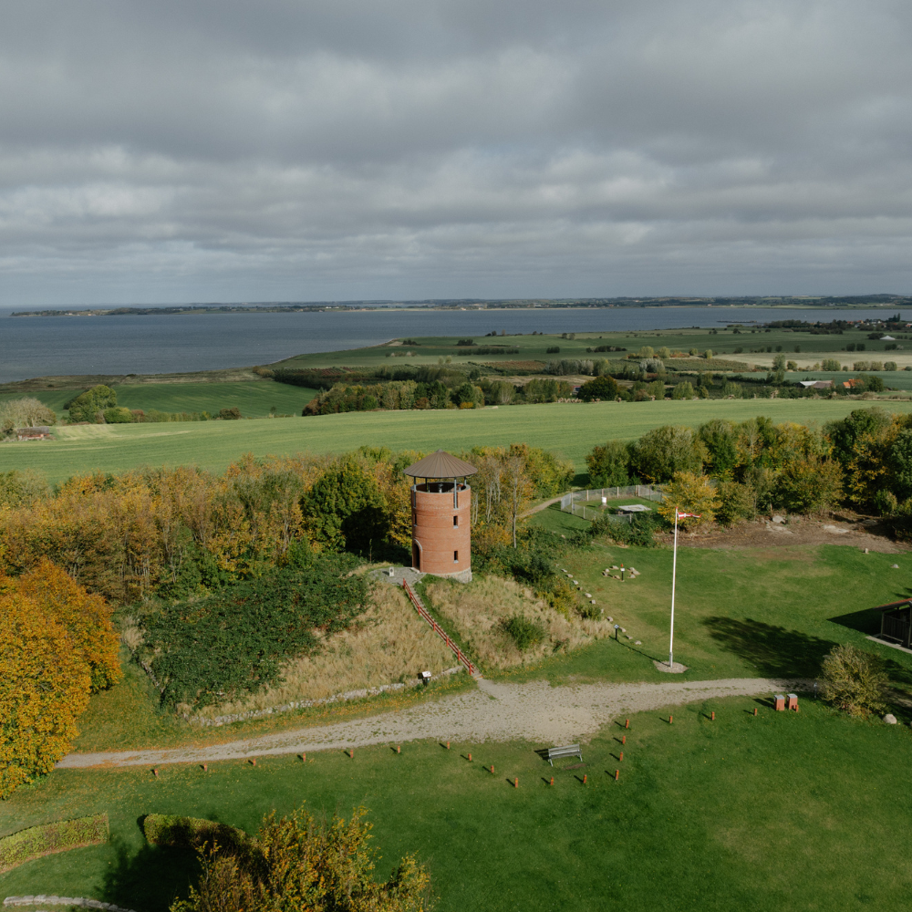 Munke Bakke set fra luften, hvor udkigstårnet er placeret i midten med mark, skov, hegn og havet bagved 