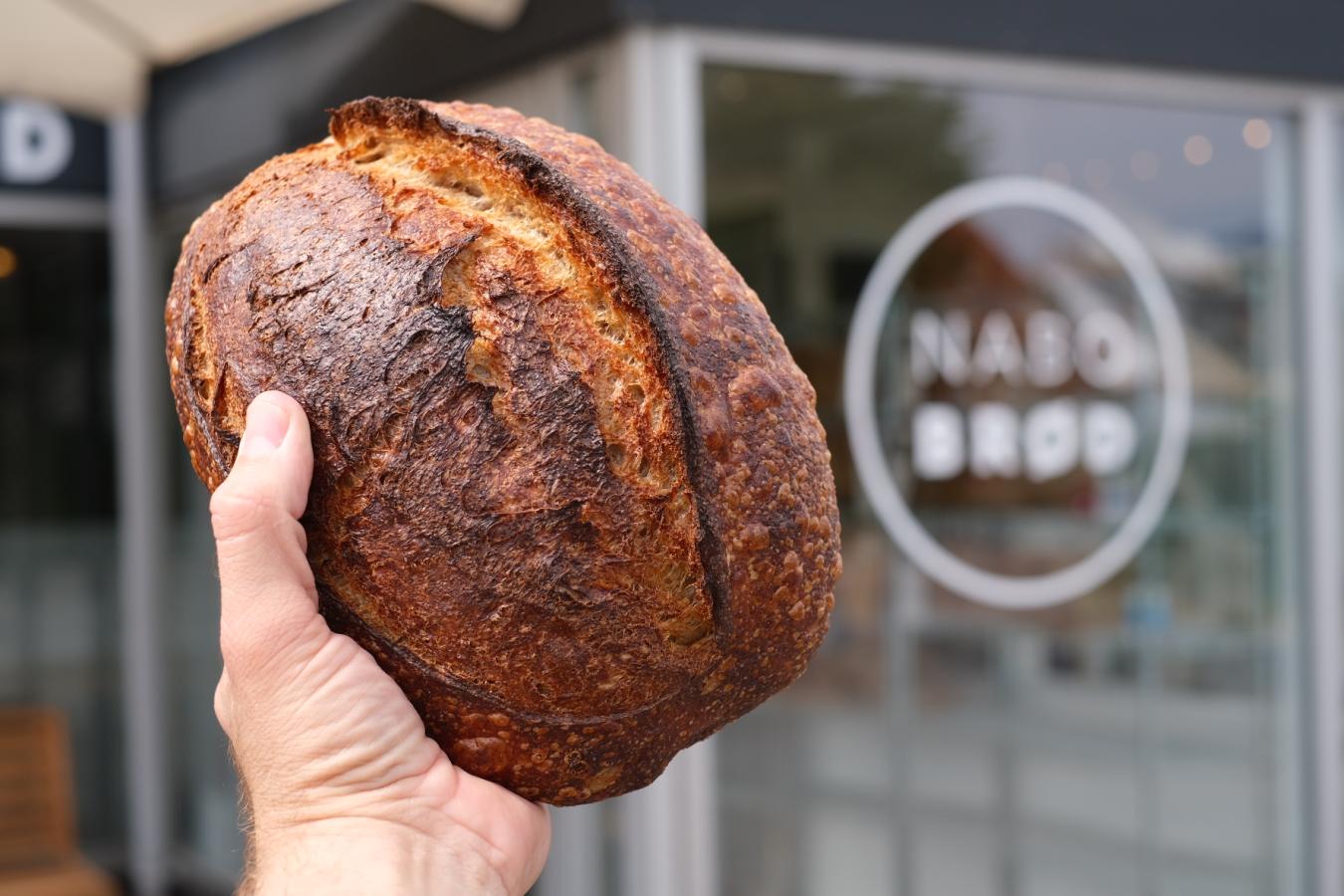 Et mørkebrunt franskbrød holdes i hånden op foran facaden på en butik, der hedder nabobrød. 