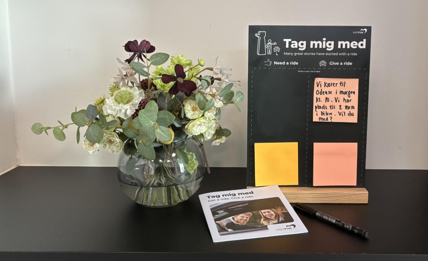 Tag-mig-med-tavle på bord med folder og blomster ved siden af