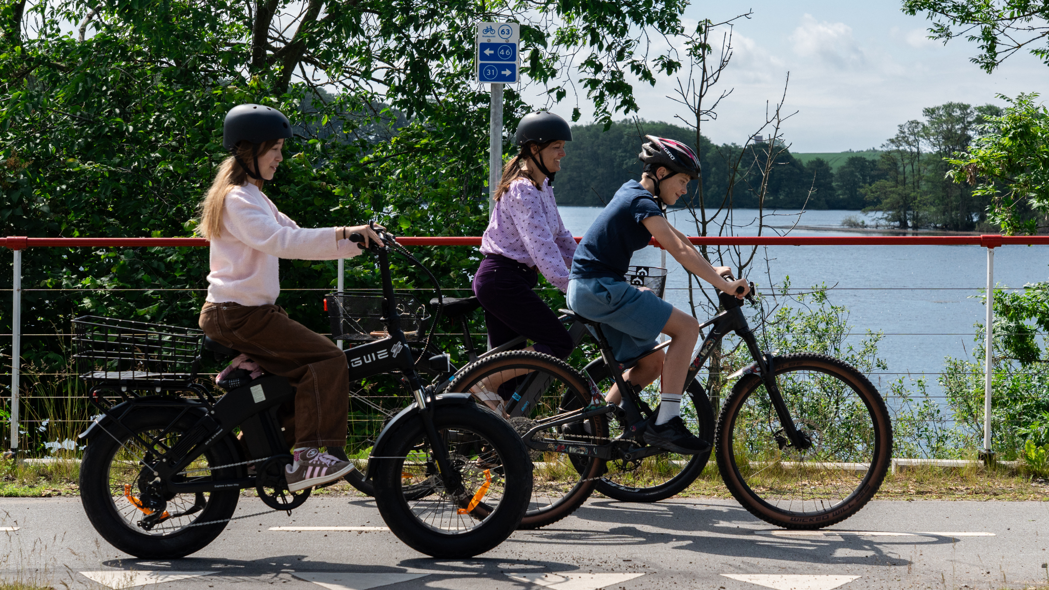 En kvinde og to børn cykler på en cykelsti lige ud til en sø en sommerdag