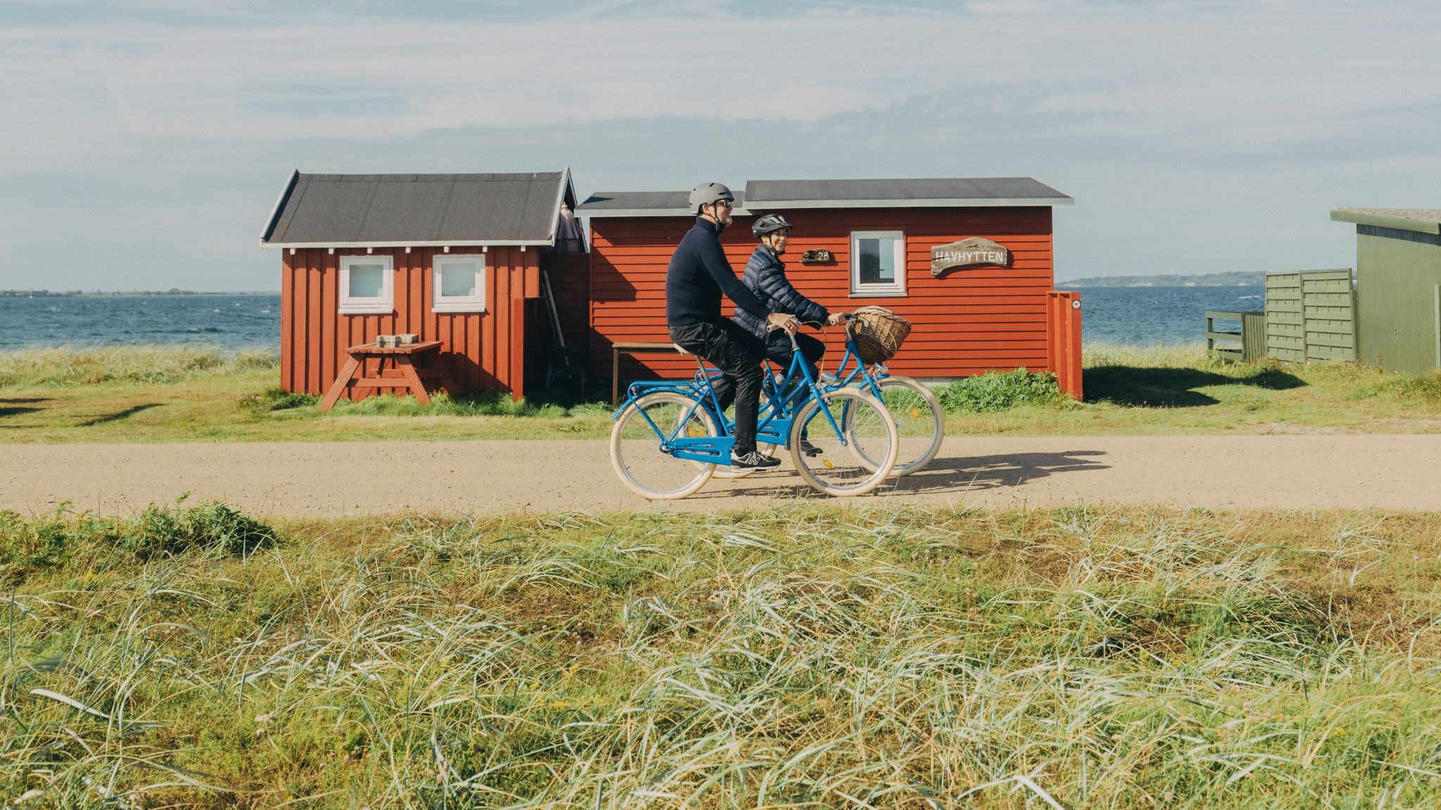 To cyklister cykler på en grusvej med havet og små badehuse bagved. 
