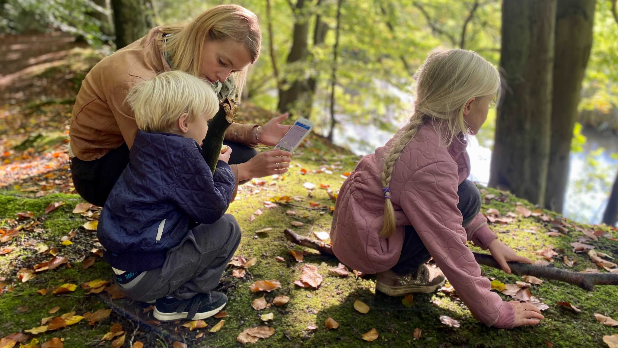 To lyshårede børn sidder på hug på en skovbund, mens deres mor holder en telefon og læser højt fra eventyr.