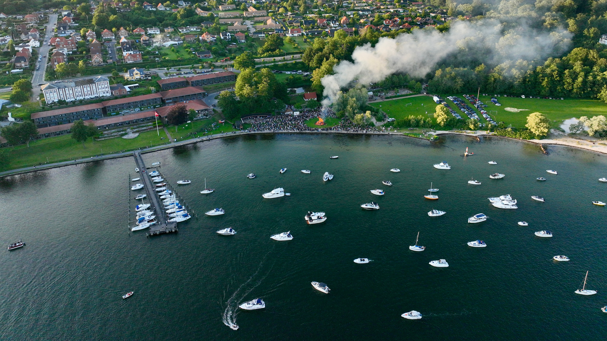 Dronefoto af et Skt Hans bål på en græseng lige ud til kyst og hav, hvor 20-30 både lægger for anker