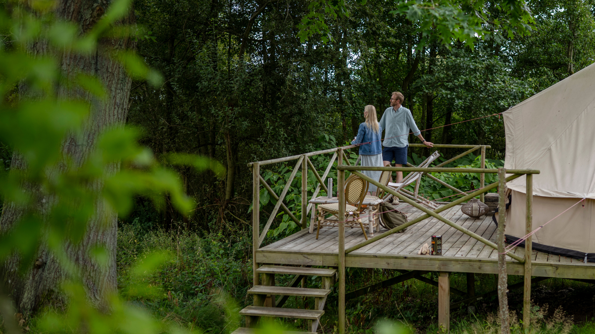 Et par står på et trædæk foran et glampingtelt, med naturen som baggrund.
