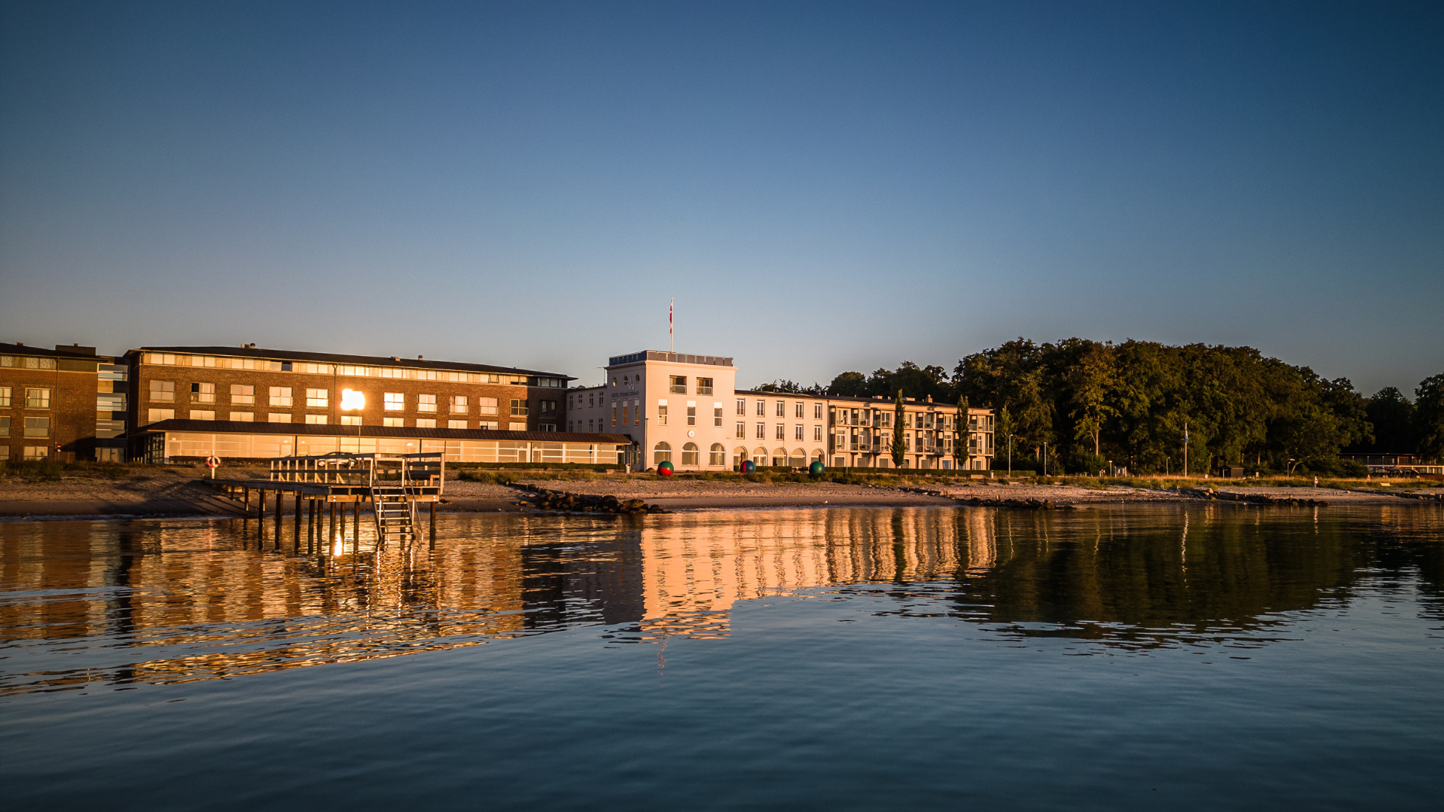 Hotelfacade lands en kystlinje i solopgangslyst med roligt vand foran, grøn skov ved siden af.