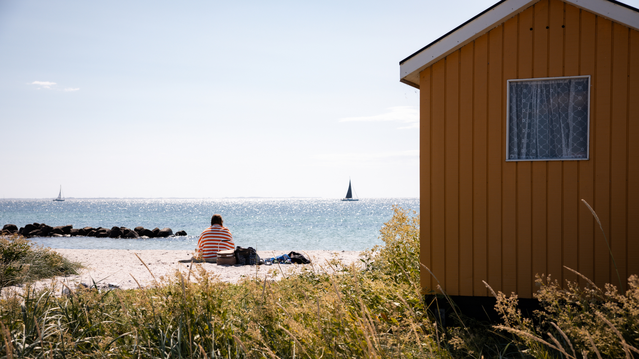 Et gult badehus på en hvid sandstrand med en person siddende på stranden ud til havet, hvor der sejler sejlskibe en sommerdag