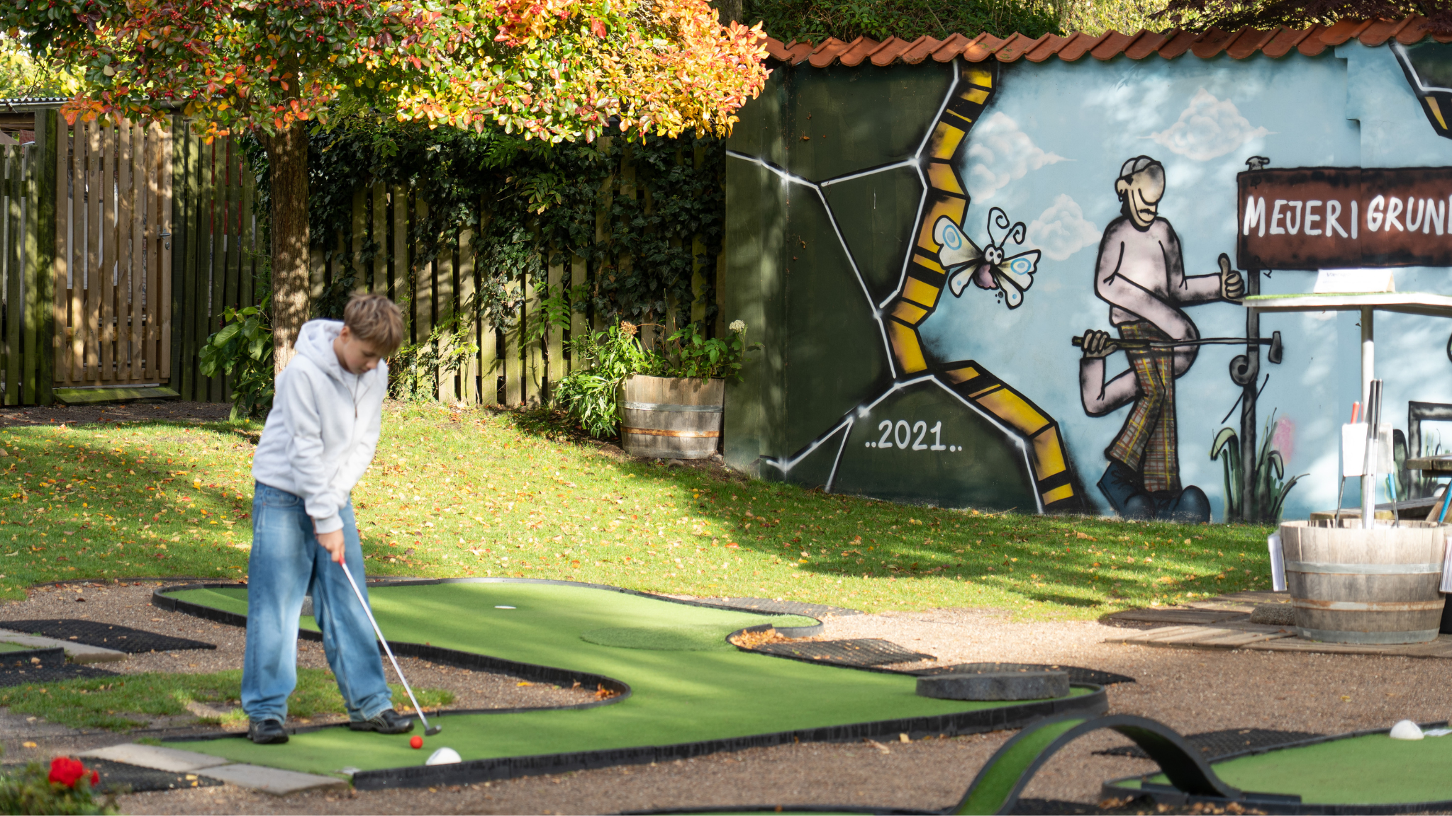 En ung dreng står med en golfkølle på en minigolfbane med græsplæne og træer omkring sig og en væg med graffiti på.