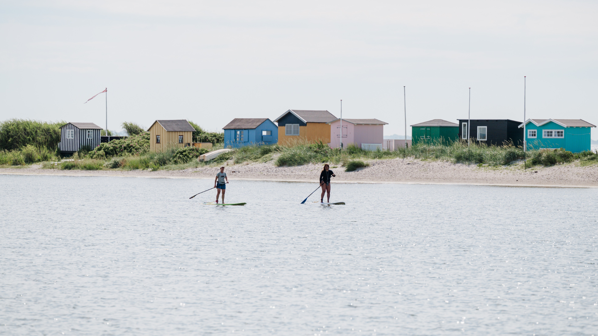 To kvinder står på stand up paddle boards på vand foran en landtange med farverige badehuse