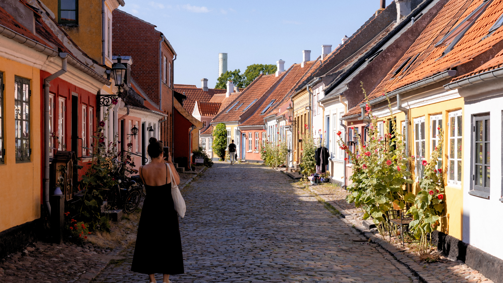 En kvinde går ned ad en brostensbelagt gade, omgivet af bygninger og grønne planter.