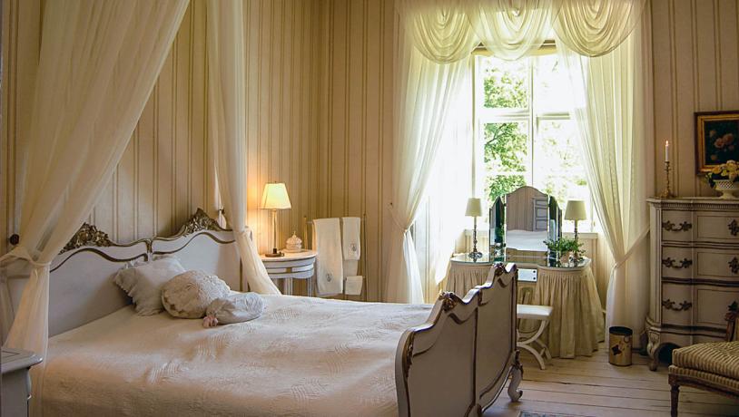 Suite med smuk, gammel seng, oplyste hvide gardiner og andet gammelt interiør på på Broholm Slot.