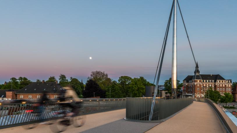 Cyklist på Byens Bro i Odense. I baggrunden ses smukke farver på aftenhimmelen og byens huse og tage.