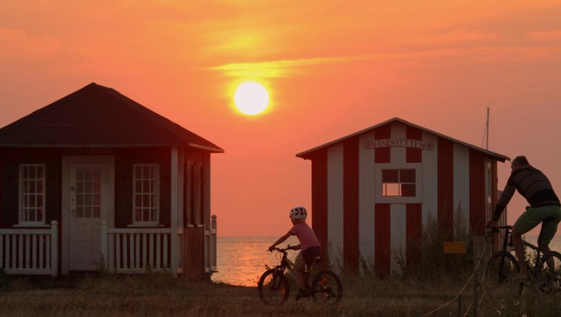 Far og barn på cykel i knaldrød solnedgang foran to små badehuse på Ærø.