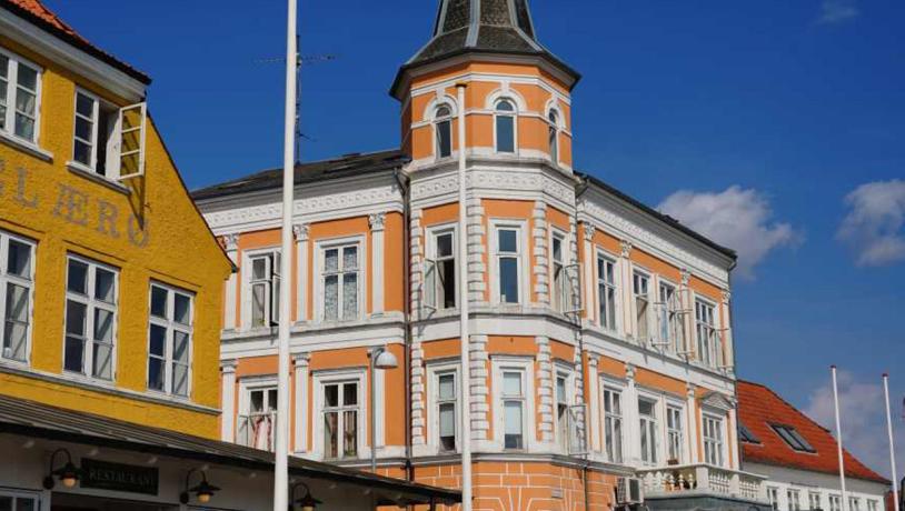 I venstre side ses toppen af en sennepsgul bygning med teksten Hotel Ærø. I højre side ses en orange bygning med hvide vinduesrammer og et tårn.