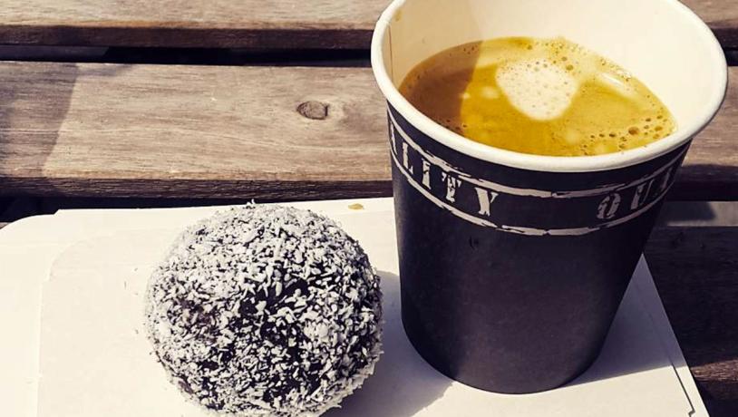 En kop kaffe og en romkugle med krymmel serveret på paptallerken på et træbord.