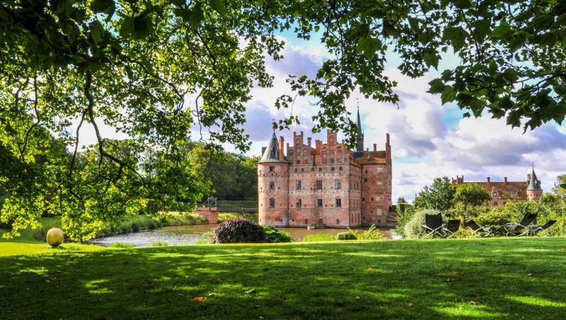 Egeskov slot med grønt græs i forgrunden og grønne træer og blå himmel i baggrunden.