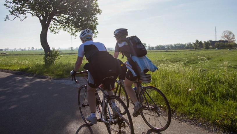 To cyklister i fart hen ad vejen. Træ og grønne marker i baggrunden.