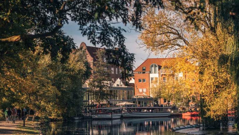 Odense Aafart på en solskinsdag i efteråret og folk går tur på stierne. Man kan se to både og vandcykler, som ligger i vandet foran Nelles bar. 