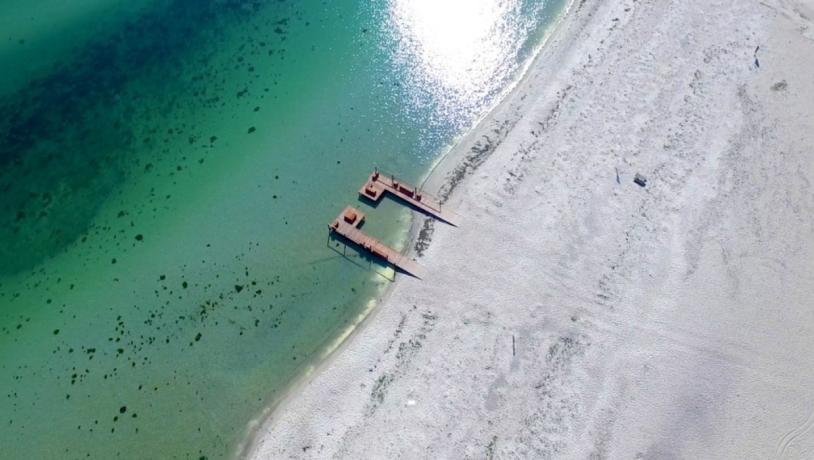 Strand set ovenfra med lyst sand og grønligt vand.