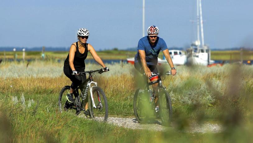 To cyklister på grussti i grønt landskab med to hvide både i baggrunden.