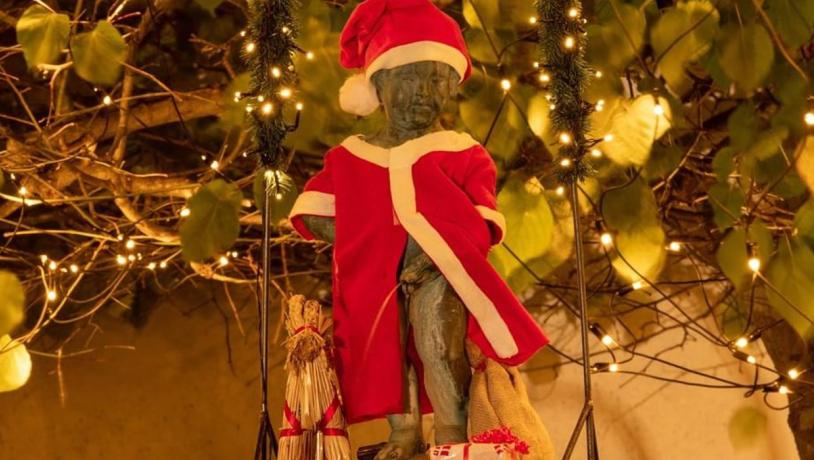 Statuen Mannekin Pis i Bogense klædt i julekostume. I baggrunde ses en lyskæde med lys i og grønne blade.