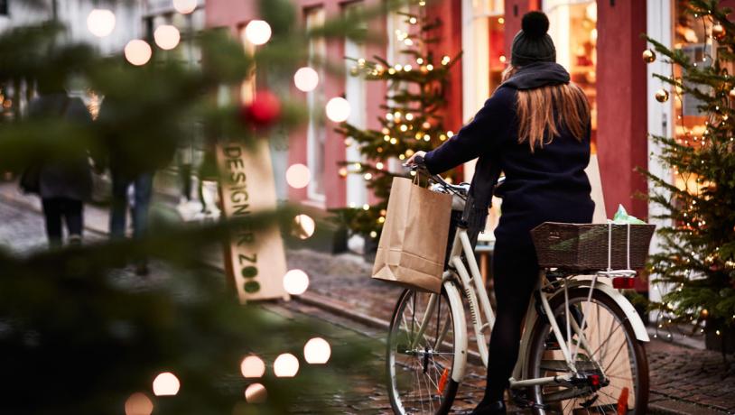 I venstre side er slørede juletræsgrene med julelys. I højre side cykler en kvinde med hue og halstørklæde. Hun har en pose i hånden og i hendes cykelkurv bagpå ligger endnu en pose. Bag hende ses en rød bygning og to små juletræer med guldkugler og lyskæder.