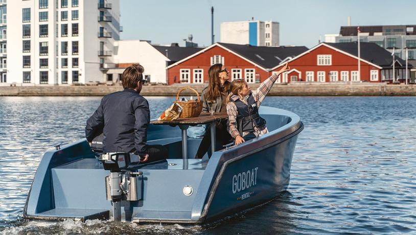 GoBoat-båd på vandet med tre personer i. Den ene peger på noget, som de andre kigger efter. I baggrunden ses to røde pakhuse og stor hvid bygning.