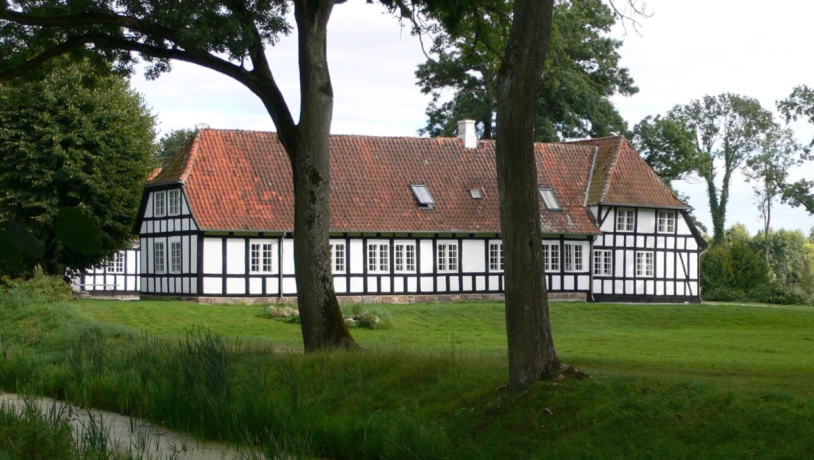 En herregård i hvidt bindingsværk med rødt tag, med et stort træ foran, der fremhæver ejendommens charme og naturlige omgivelser.