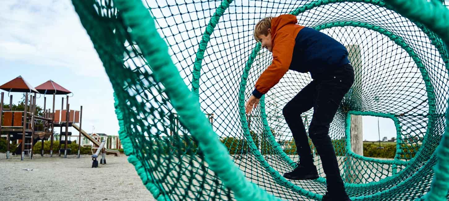 Et større barn inde i 'fiskeruse' på en legeplads med faldsand. I baggrunden ses et legetårn.