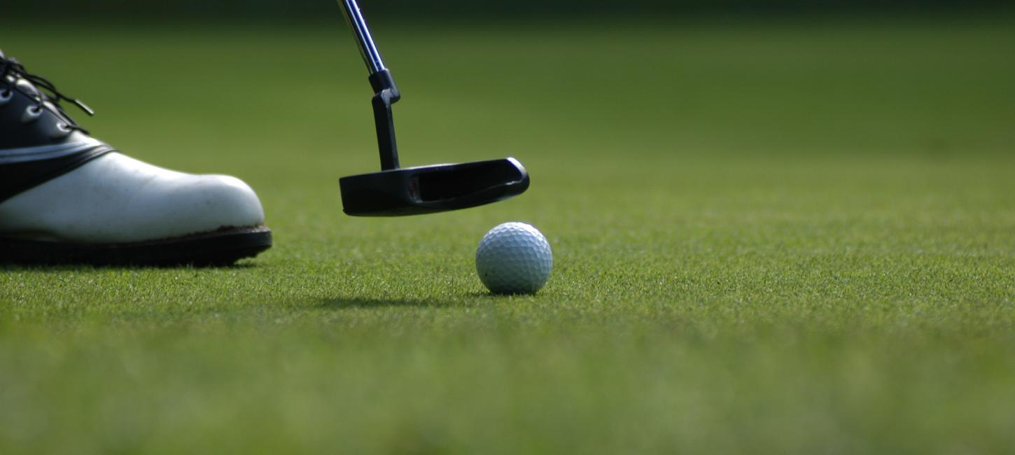 Nærbillede af golfbold på fairway. Golfkølle nærmer sig bolden og det forreste af en hvid golfsko ses til venstre i billedet.