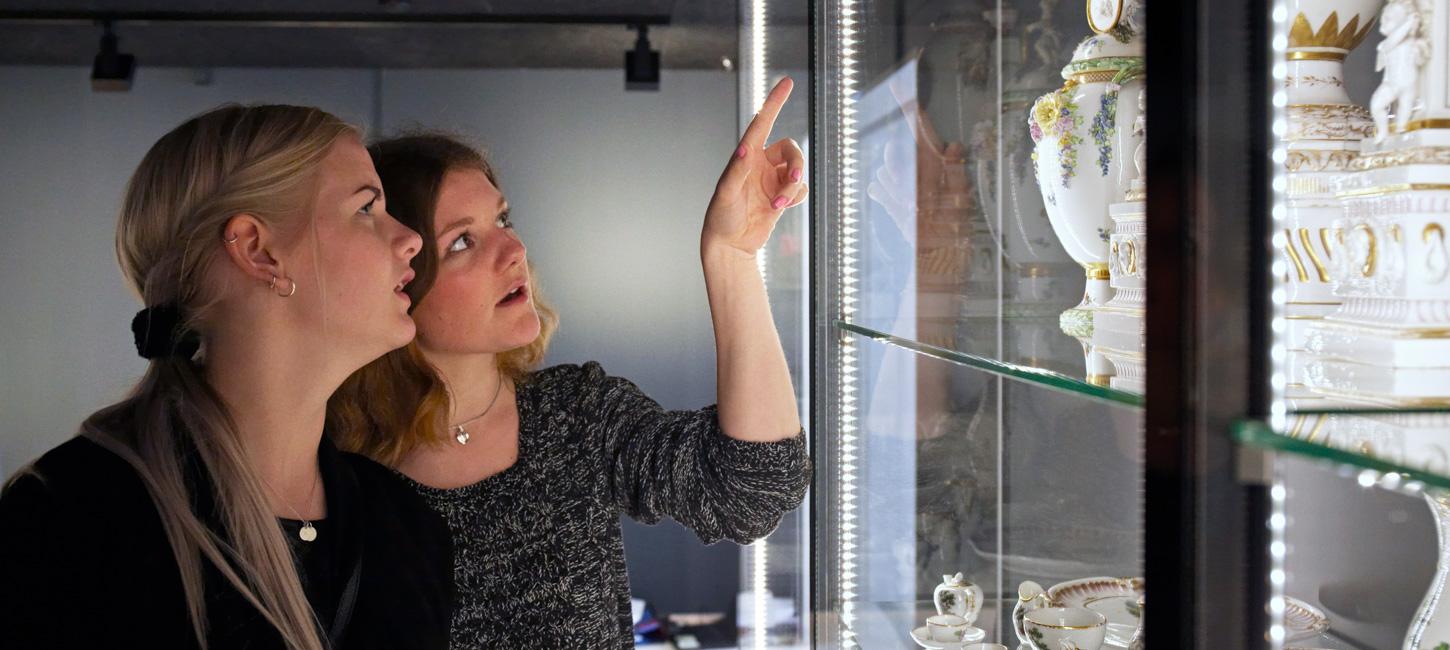 To kvinder står foran en glasmontre. Den ene peger ind i glasmontren. I glasmontren står hvidt porcelæn pyntet med guld detaljer.