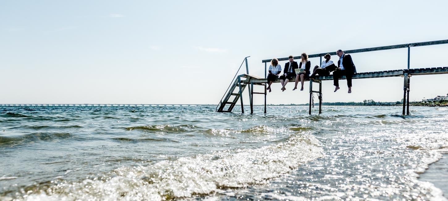 En lille gruppe forretningsfolk sidder på en badebro og kigger ud over vandet.