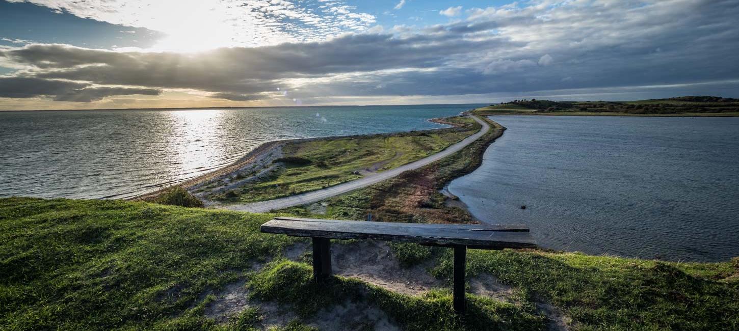 Udsigt over tange med smal vej og vand på begge sider. Solen stråler dramatisk gennem lavthængende grå skyer. I forgrunden står en træbænk.