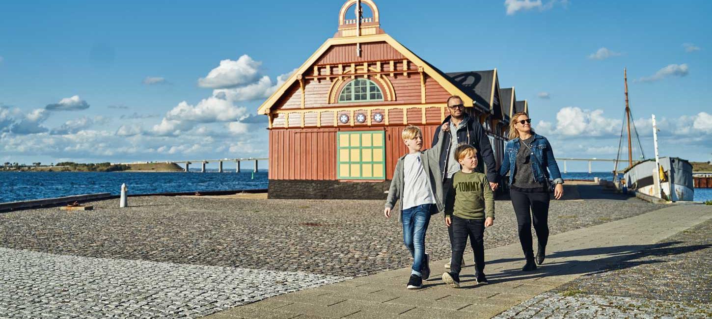 To børn og to voksne går tur ved en havn, hvor man bagved kan se et pakhus i gule og røde farver. 