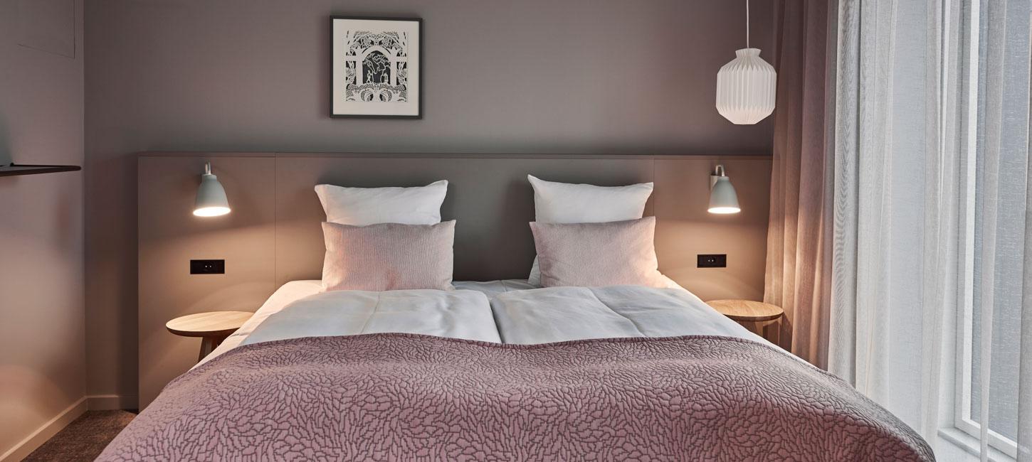 En dobbelt seng med lyserødt sengetæppe og pyntepuder. På hver side er der et sengebord af træ og en tændt læselampe. Over sengen hænger et billede på en grå væg.