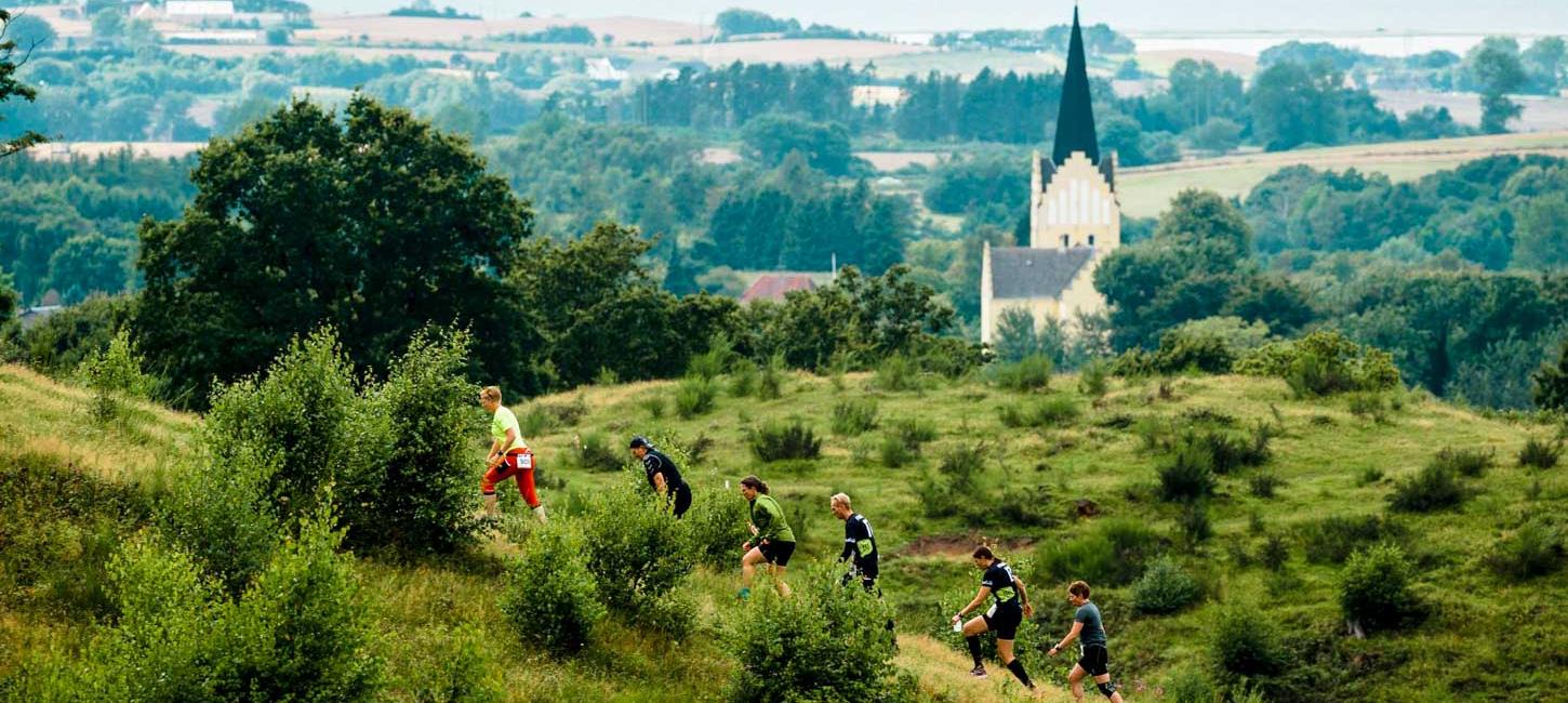 Seks mennesker i løbetøj ses løbe op ad en stejl bakke ved Svanninge Bakker. Bag ved dem ligger en hvid kirke med et højt spir.