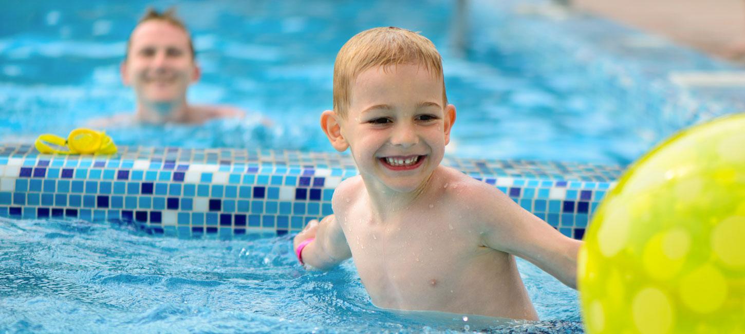 En glad dreng med et stort smil svømmer i en pool med blå mosaik-fliser. Bag ham anes en voksen, der smilende ser på barnet. En del af en stor grøn badebold er synlig i forgrunden.